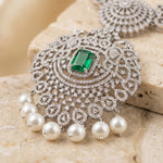 Silver-toned ornate pendant necklace with a rectangular green emerald-cut center stone and dangling white pearl beads, displayed on a beige stone surface.