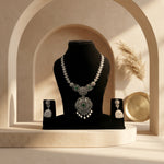 An ornate silver-toned jewelry set displayed on a black mannequin bust, including a necklace, earrings, and matching holder pieces, with a green stone accent in the center. The set is styled on a beige platform with an arched backdrop and soft warm lighting.