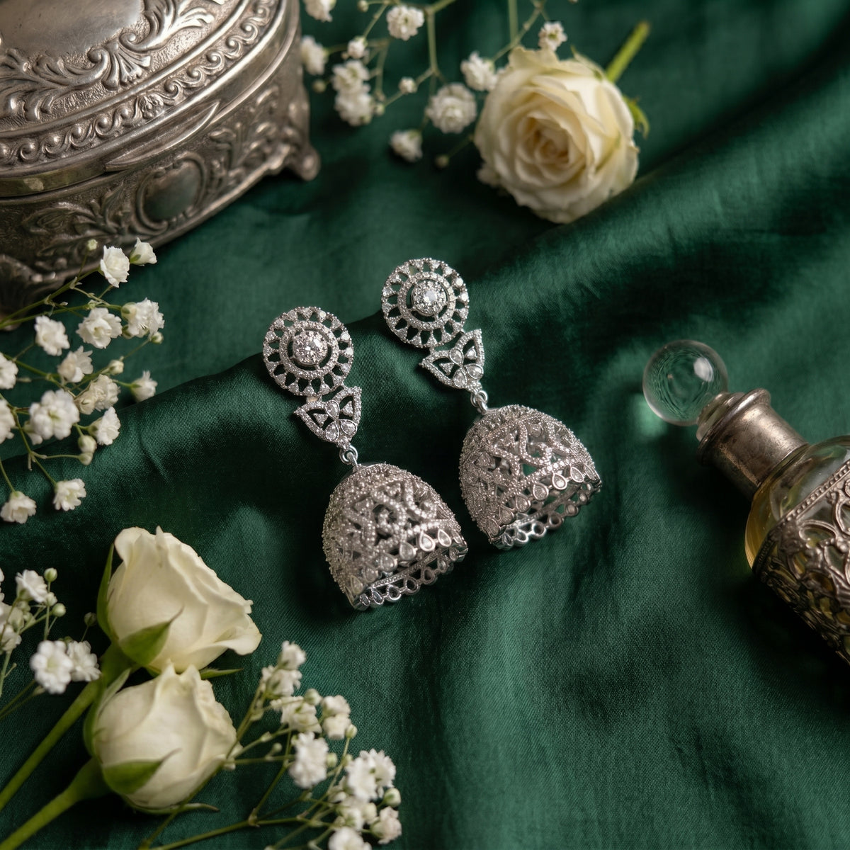 A pair of silver-toned filigree jhumka earrings with circular floral motifs, displayed on a dark green fabric background with white roses, baby's breath, and an ornate silver container.
