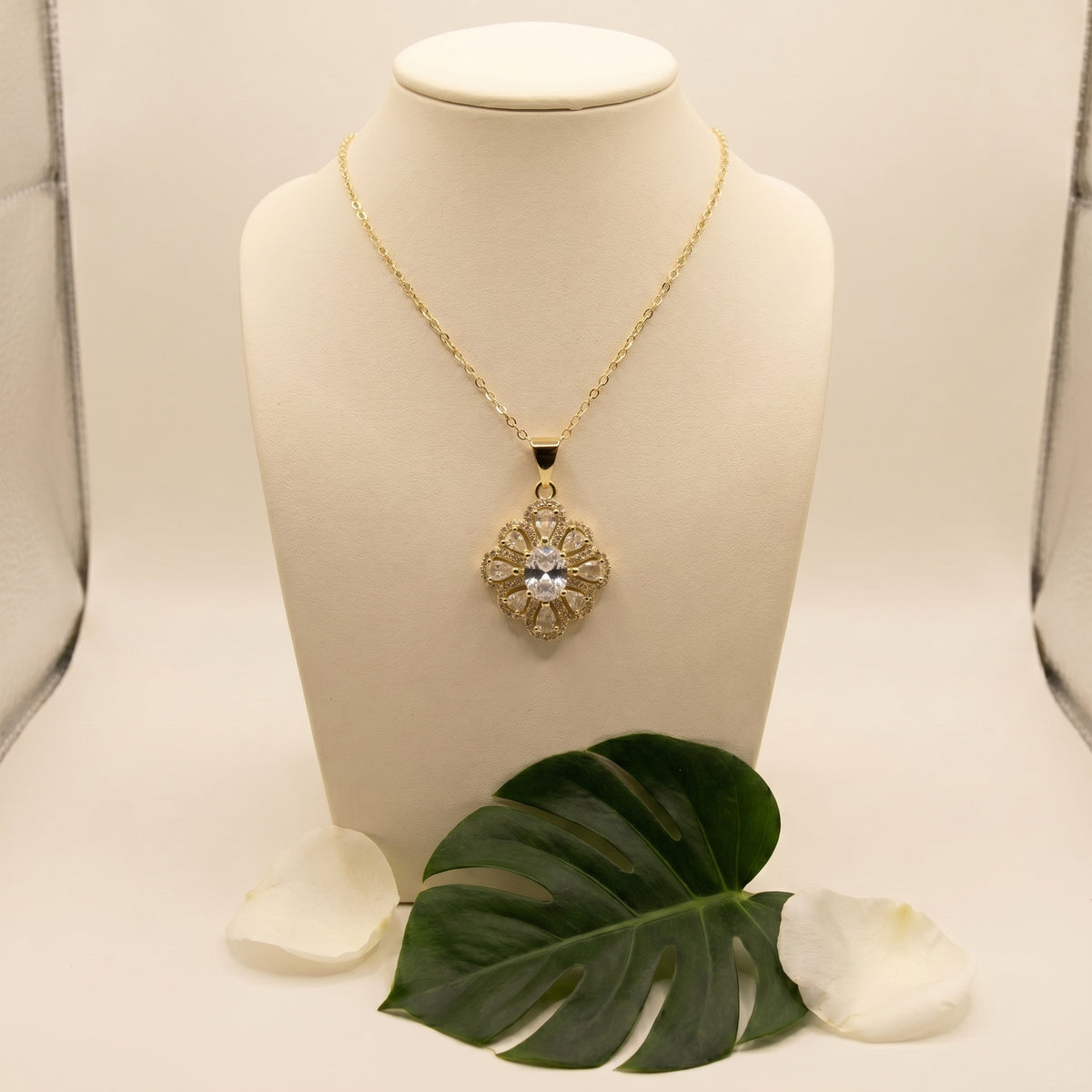 A gold pendant necklace displayed on a cream jewelry bust, featuring a floral starburst design with a central clear gemstone. A green monstera leaf and white petals are placed at the base of the bust against a light beige background.