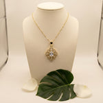A gold pendant necklace displayed on a cream jewelry bust, featuring a floral starburst design with a central clear gemstone. A green monstera leaf and white petals are placed at the base of the bust against a light beige background.