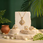 A gold jewelry set displayed on stone pedestals, featuring a necklace, earrings, and rings with clear floral-shaped stones, styled with green leaves, white tulips, and a terracotta pot against a dark green background.