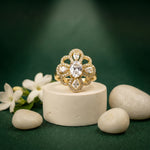 A gold ring with a floral, scalloped setting and clear teardrop gemstones, displayed on a small white pedestal with decorative white flowers and smooth stones against a dark green background.