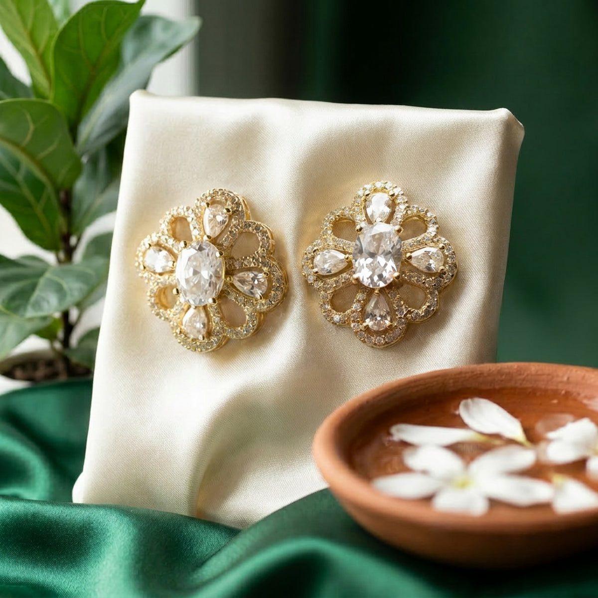 A pair of ornate gold-toned brooch-style earrings with clear teardrop and oval stones, displayed on a cream satin stand with green fabric, a plant, and a small bowl of petals in the background.