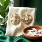 A pair of ornate gold-toned brooch-style earrings with clear teardrop and oval stones, displayed on a cream satin stand with green fabric, a plant, and a small bowl of petals in the background.