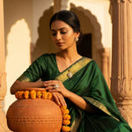 A woman wearing a dark green saree with a gold border sits beside a terracotta pot decorated with orange marigold garlands, set against a warm architectural background with arches.