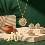 Gold jewelry set featuring a pendant necklace, matching earrings, and a ring, all adorned with clear crystal-like stones in a sunburst design, displayed on textured stone blocks with a small terracotta pot and white flower buds against a dark green background.