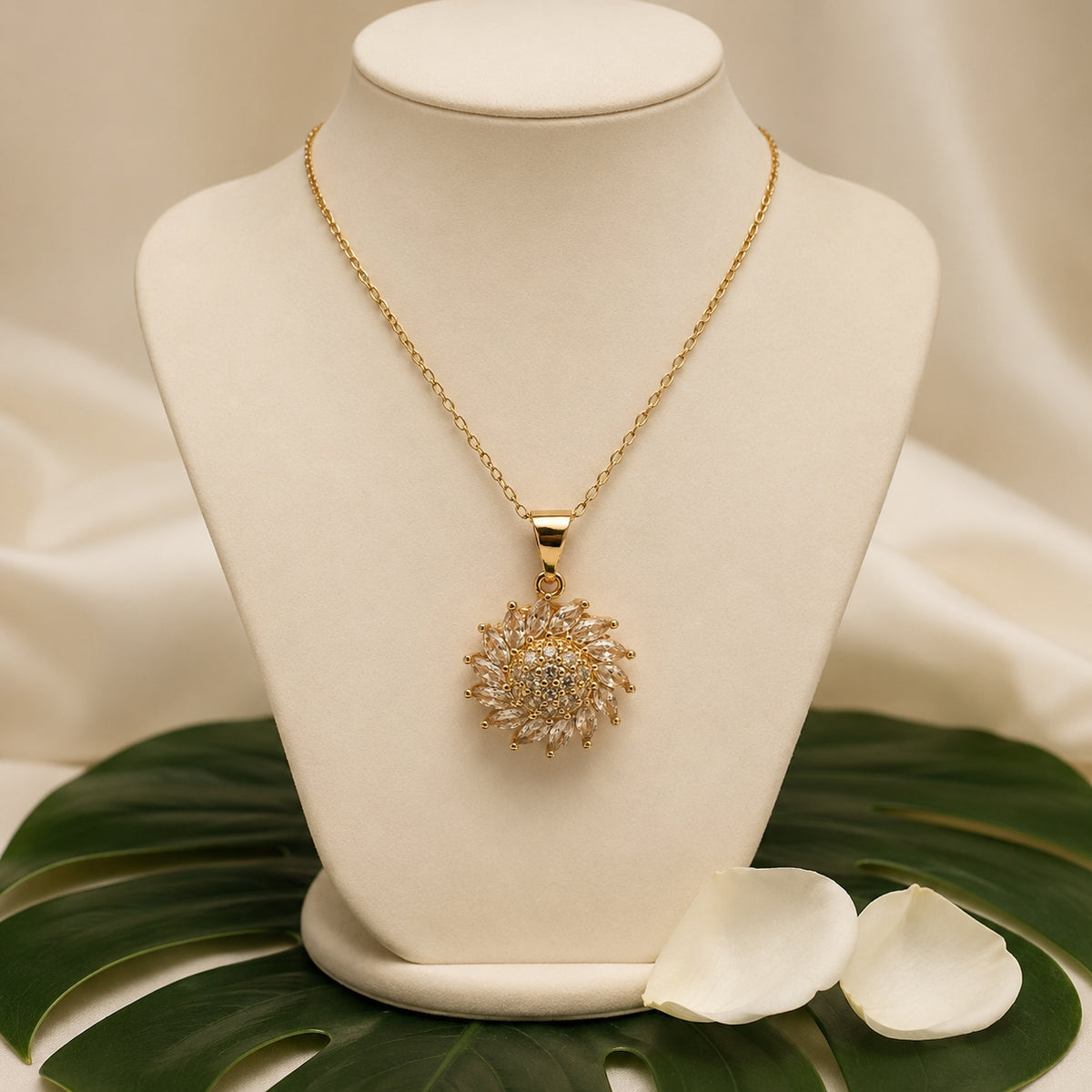 A gold chain necklace with a round sunburst pendant set with clear stones, displayed on a cream jewelry bust. The bust is styled with a green monstera leaf and white petals on a draped beige satin background.