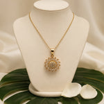 A gold chain necklace with a round sunburst pendant set with clear stones, displayed on a cream jewelry bust. The bust is styled with a green monstera leaf and white petals on a draped beige satin background.