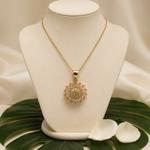 A gold pendant necklace with a sunburst design set with clear stones, displayed on a cream-colored jewelry bust. The bust is styled with a green monstera leaf and white petals on a draped cream satin background.