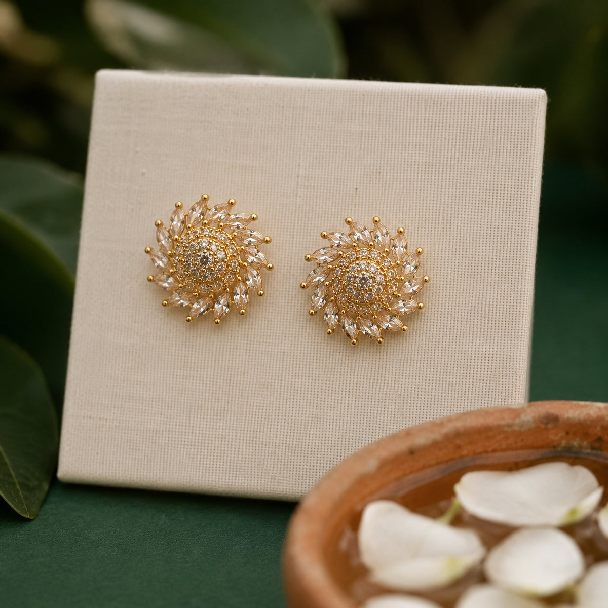 A pair of gold and crystal sunburst stud earrings displayed on a cream-colored textured card, set against a dark green background with blurred leaves and a terracotta bowl of petals.