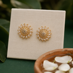 A pair of gold-toned stud earrings with clear rhinestone-like accents, displayed on a cream fabric card. The card is set against a dark green background with blurred leaves and a terracotta bowl of petals in the foreground.