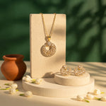 Gold jewelry set featuring a circular pendant necklace and matching stud earrings, displayed on cream-colored stone pedestals with a green leafy backdrop and small white flower buds.