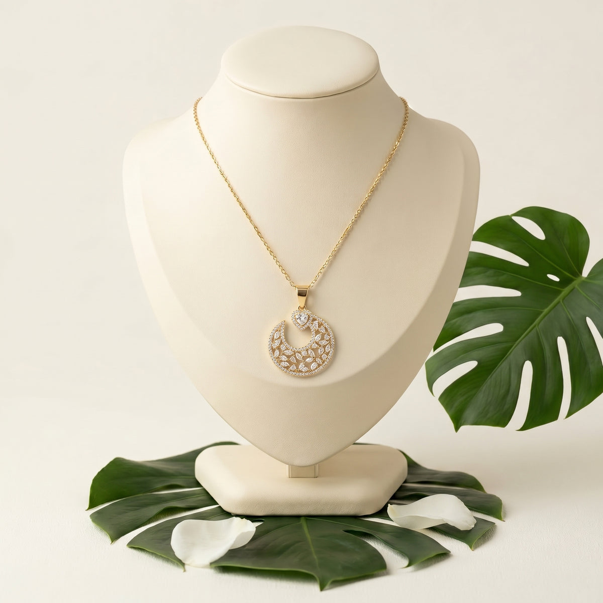 A gold-tone necklace with a circular pendant set with clear stones, displayed on a cream jewelry bust with green tropical leaves on a light beige background.