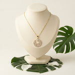 A gold-tone necklace with a circular pendant set with clear stones, displayed on a cream jewelry bust with green tropical leaves on a light beige background.