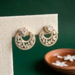 A pair of gold crescent-shaped stud earrings adorned with clear pavé stones and a central round clear stone, displayed on a beige fabric stand against a dark green background with a small wooden dish in the foreground.