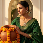 A woman in a dark green saree with gold zari detailing holds a terracotta pot decorated with orange and yellow marigold garlands. She is wearing a gold pendant necklace and matching earrings, set against a warm, sunlit architectural background with an arched doorway.