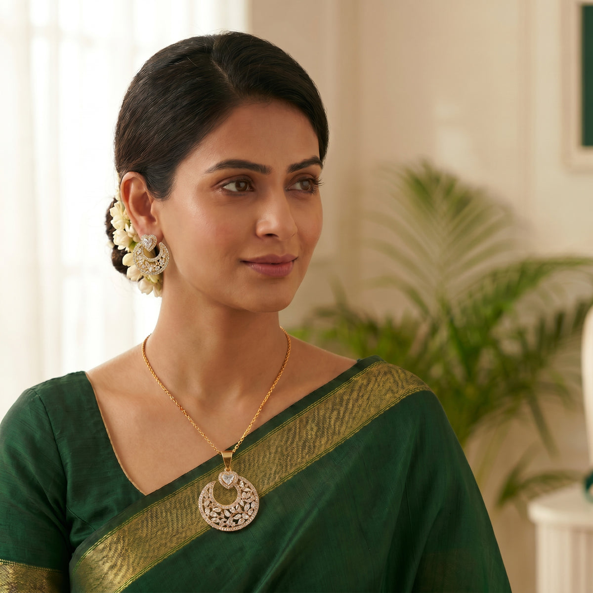 A woman wearing a dark green saree with a gold border, a gold necklace with a circular silver-toned pendant, and white floral earrings, posed in a softly lit indoor setting.