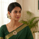 A woman wearing a dark green saree with a gold border, a gold necklace with a circular silver-toned pendant, and white floral earrings, posed in a softly lit indoor setting.